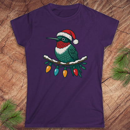 A purple short-sleeve crewneck t-shirt featuring a graphic of a hummingbird wearing a red Santa hat and perched on a branch with Christmas lights, displayed flat on a rustic wood background with pine needles.