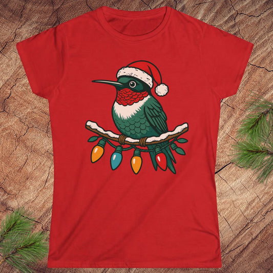 A red short-sleeve crewneck t-shirt featuring a graphic of a green hummingbird wearing a red and white Santa hat, perched on a branch with colorful Christmas lights, laid flat on a rustic wood background with pine needles.