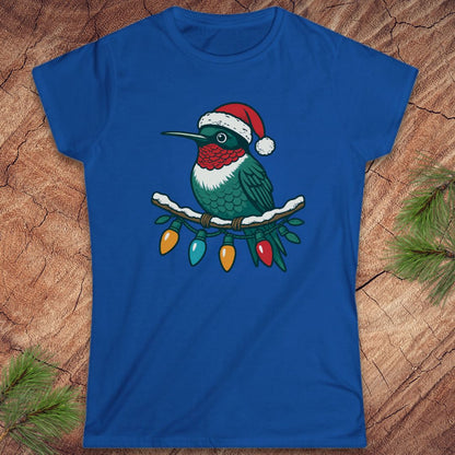 A royal blue short-sleeve t-shirt featuring a graphic of a green hummingbird wearing a red Santa hat and perched on a branch with colorful Christmas lights, displayed flat on a rustic wood background with pine needles.