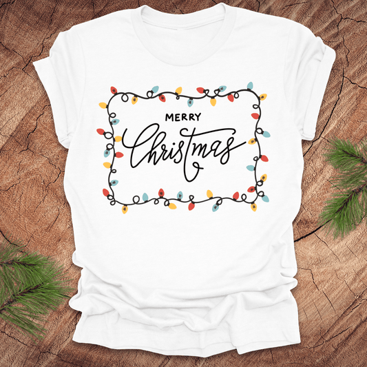 White t-shirt with christmas lights graphic and 'Merry Christmas' design on a wooden surface