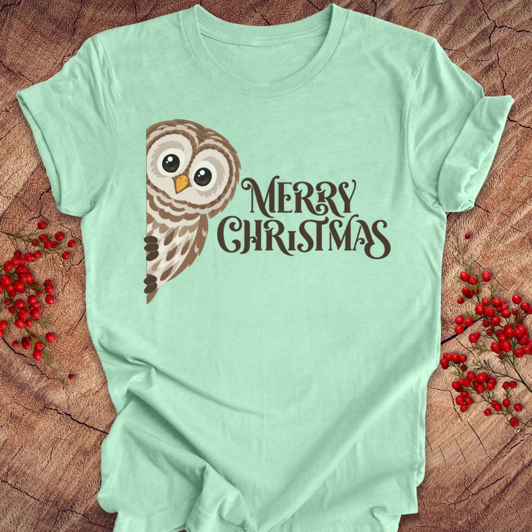 A mint green short-sleeve t-shirt with a graphic of a brown owl and the text 'MERRY CHRISTMAS' in dark brown, laid flat on a rustic wood background with red berries.