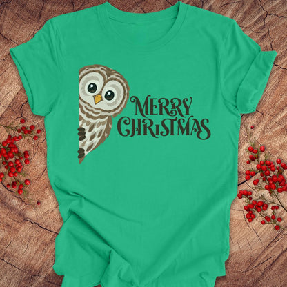 A bright green short-sleeve t-shirt with a 'Merry Christmas' graphic featuring a brown and white owl, laid flat on a rustic wood background with red berries.