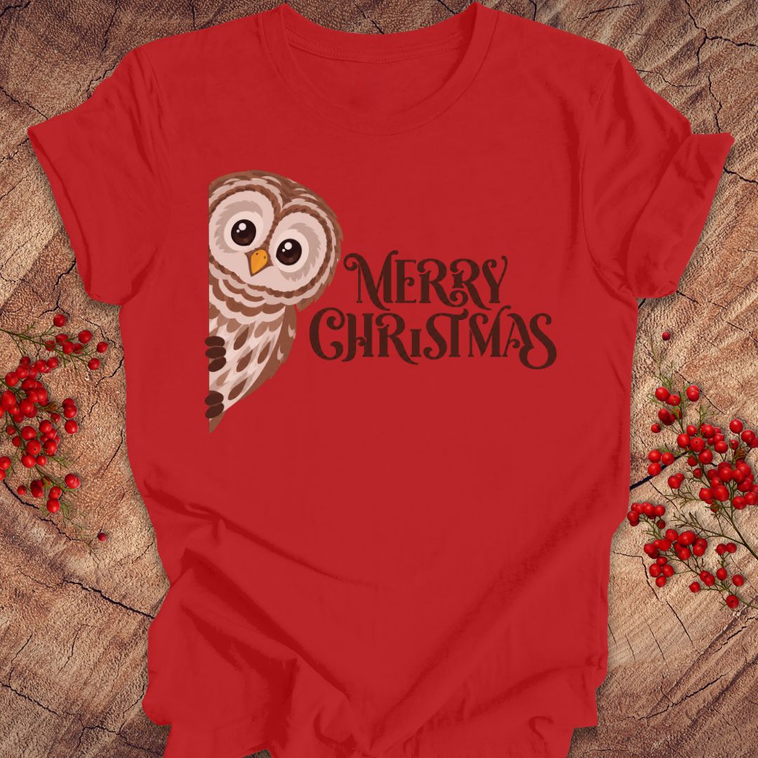 A red short-sleeve t-shirt with a graphic of a brown and white owl next to the text 'MERRY CHRISTMAS' in dark brown, laid flat on a rustic wood background with red berries.