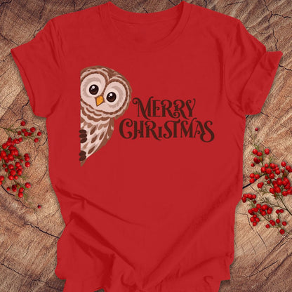 A red short-sleeve t-shirt with a graphic of a brown and white owl next to the text 'MERRY CHRISTMAS' in dark brown, laid flat on a rustic wood background with red berries.