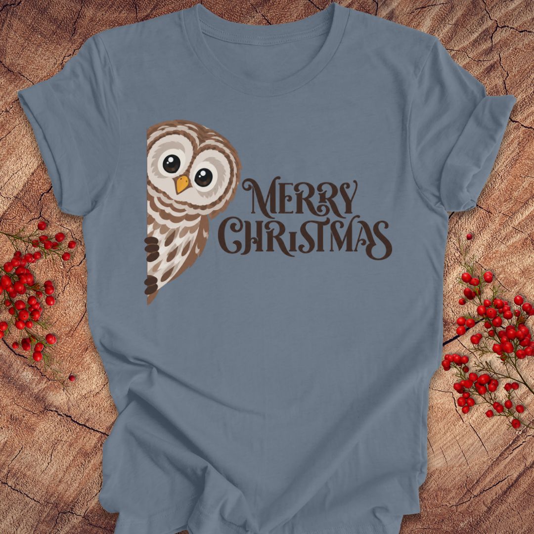 A slate blue short-sleeve t-shirt with a brown and beige owl graphic and the words 'MERRY CHRISTMAS' in dark brown script, laid flat on a rustic wood background with red berries.