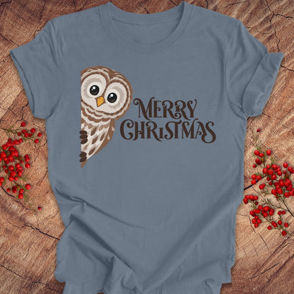 A slate blue short-sleeve t-shirt with a brown and beige owl graphic and the words 'MERRY CHRISTMAS' in dark brown script, laid flat on a rustic wood background with red berries.