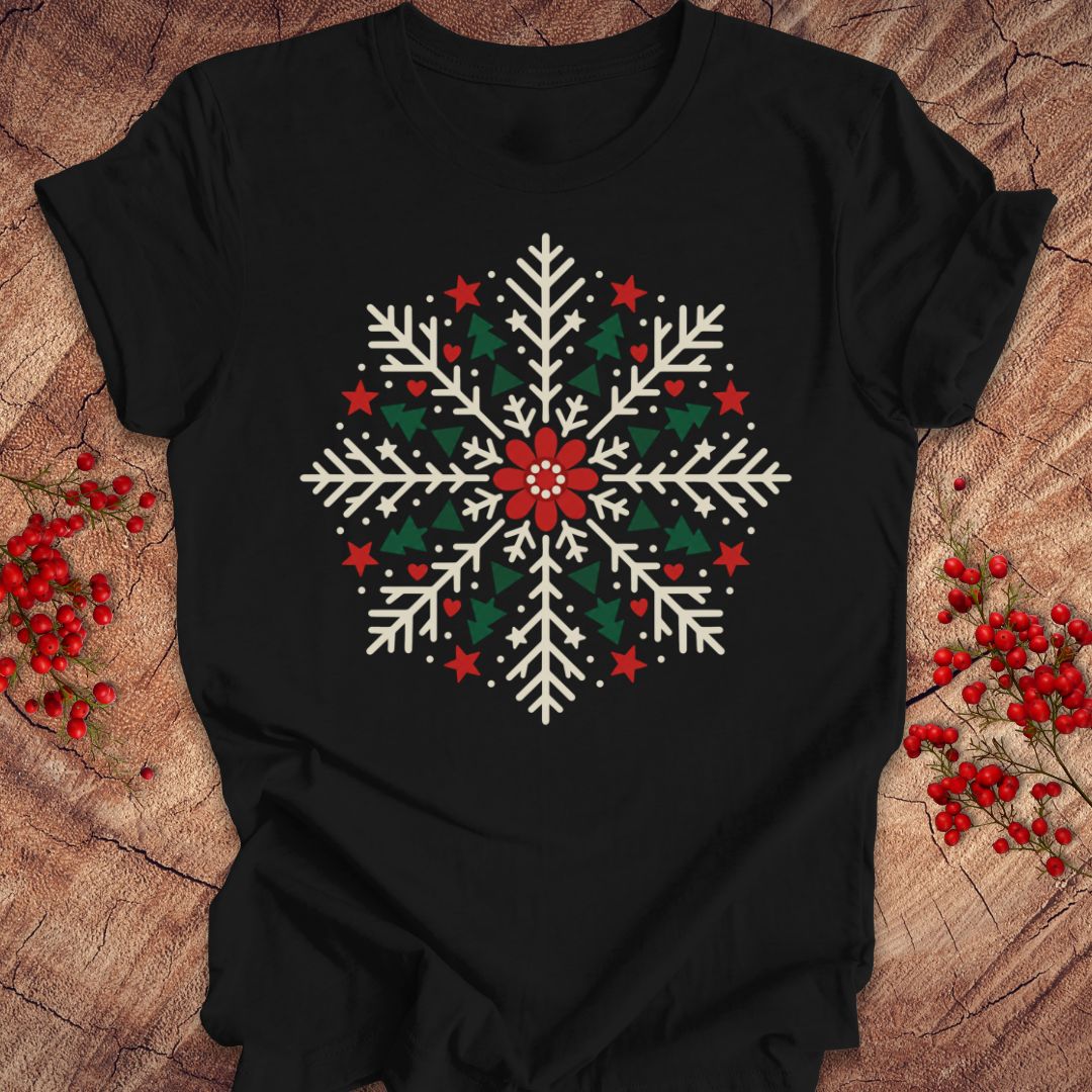 Black t-shirt with a creamy white folk-art snowflake filled with tiny green trees, red stars, and hearts around a red flower, laid flat on a rustic wood background with red winter berries.