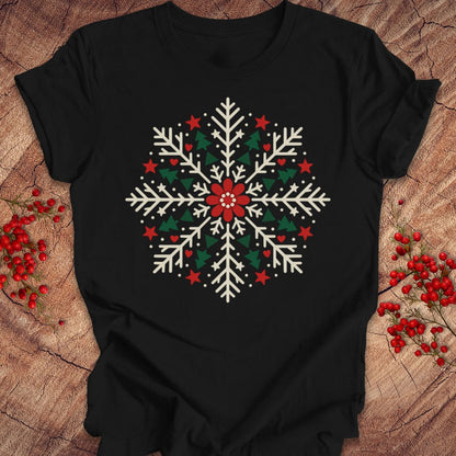 Black t-shirt with a creamy white folk-art snowflake filled with tiny green trees, red stars, and hearts around a red flower, laid flat on a rustic wood background with red winter berries.