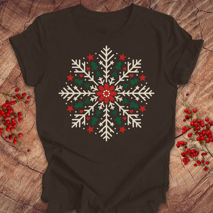 Brown t-shirt with a creamy white folk-art snowflake filled with tiny green trees, red stars, and hearts around a red flower, laid flat on a rustic wood background with red winter berries.