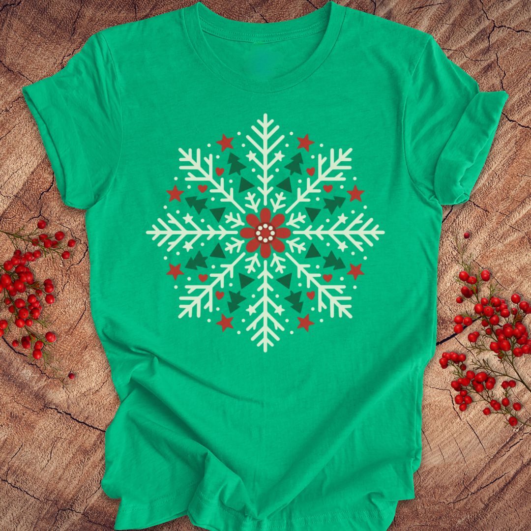 Heather kelly green t-shirt with a creamy white folk-art snowflake filled with tiny green trees, red stars, and hearts around a red flower, laid flat on a rustic wood background with red winter berries.