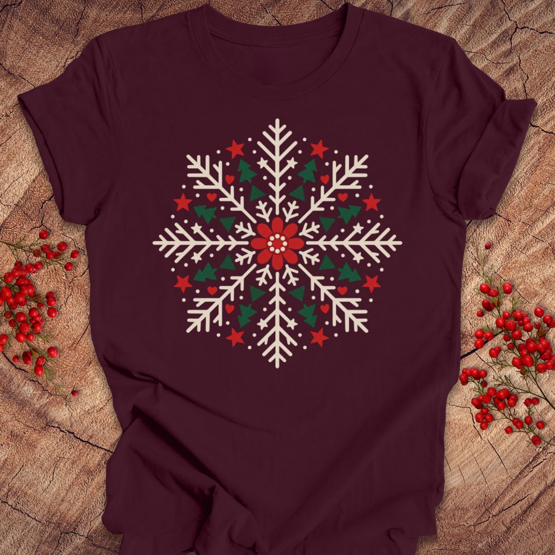 Maroon t-shirt with a creamy white folk-art snowflake filled with tiny green trees, red stars, and hearts around a red flower, laid flat on a rustic wood background with red winter berries.