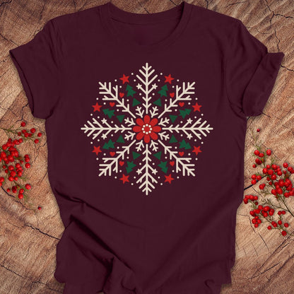 Maroon t-shirt with a creamy white folk-art snowflake filled with tiny green trees, red stars, and hearts around a red flower, laid flat on a rustic wood background with red winter berries.