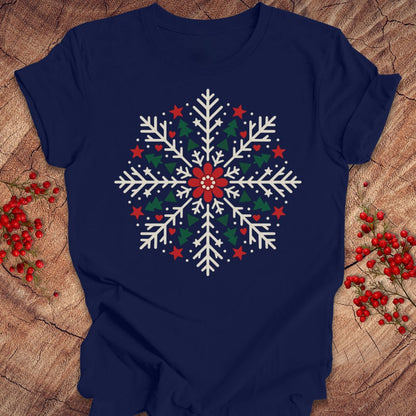 Navy t-shirt with a creamy white folk-art snowflake filled with tiny green trees, red stars, and hearts around a red flower, laid flat on a rustic wood background with red winter berries.