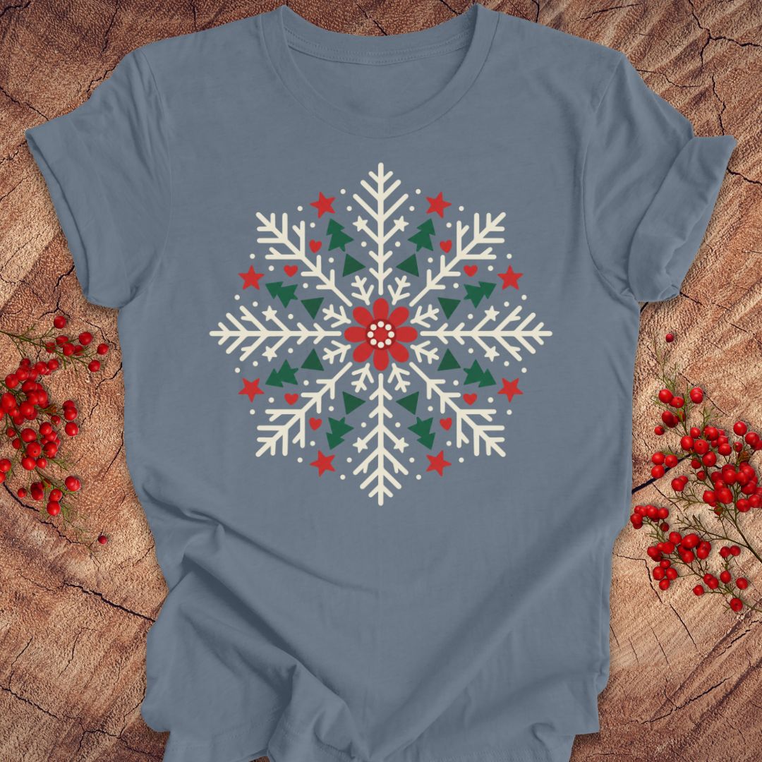 Steel blue t-shirt with a creamy white folk-art snowflake filled with tiny green trees, red stars, and hearts around a red flower, laid flat on a rustic wood background with red winter berries.