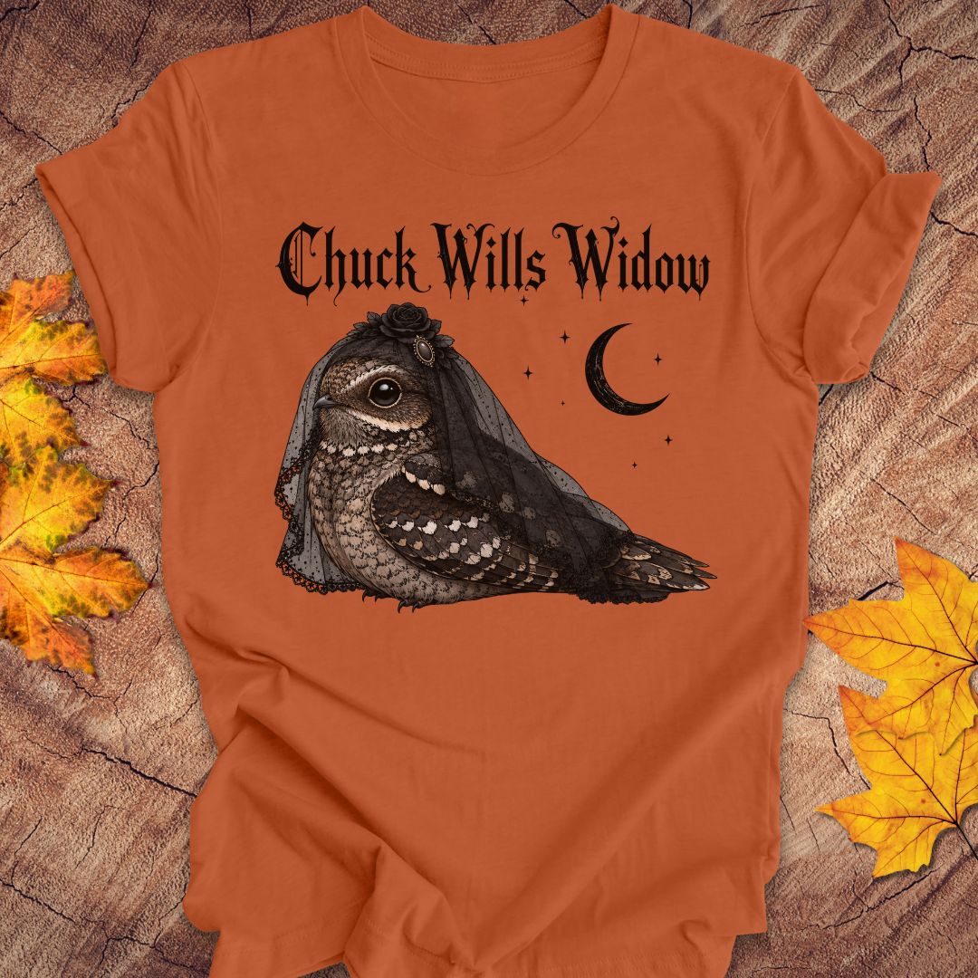 An orange crewneck t-shirt featuring a graphic of a nightjar bird wearing a black widow veil, with the text 'Chuck Wills Widow' and a crescent moon. The shirt is displayed flat on a rustic wood background decorated with autumn leaves.