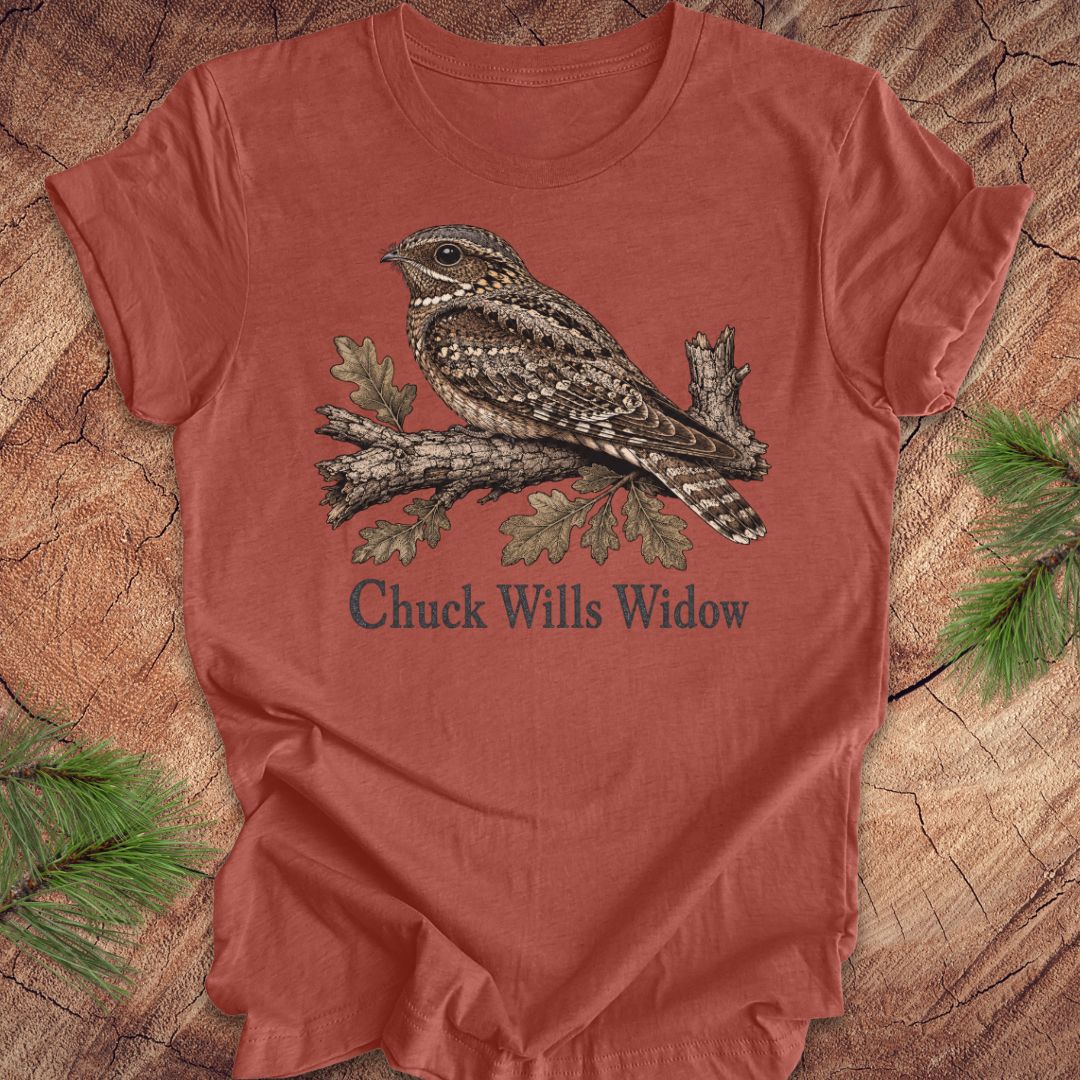A rust-colored short-sleeve t-shirt featuring a detailed graphic of a sleeping wrybill bird on a branch with the text 'Chuck Wills Widow' underneath, displayed flat on a rustic wood background with pine needles.