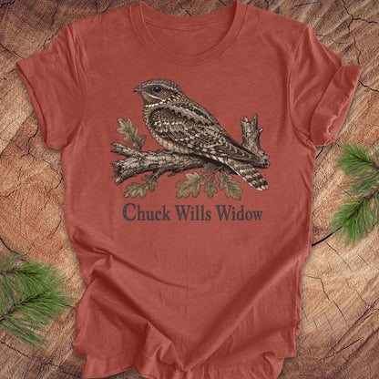 A rust-colored short-sleeve t-shirt featuring a detailed graphic of a sleeping wrybill bird on a branch with the text 'Chuck Wills Widow' underneath, displayed flat on a rustic wood background with pine needles.