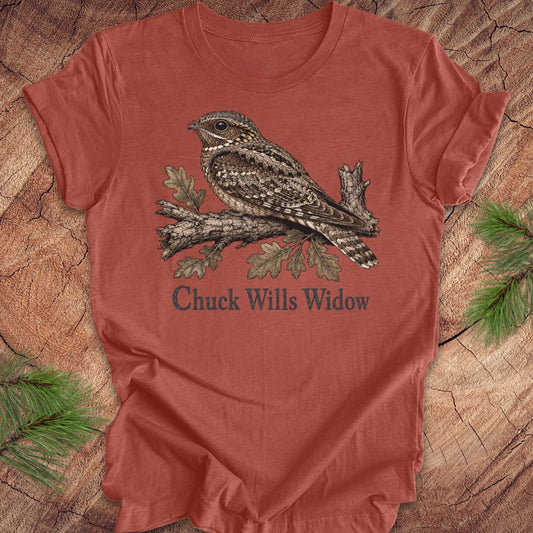 A rust-colored short-sleeve t-shirt featuring a detailed graphic of a sleeping wrybill bird on a branch with the text 'Chuck Wills Widow' underneath, displayed flat on a rustic wood background with pine needles.