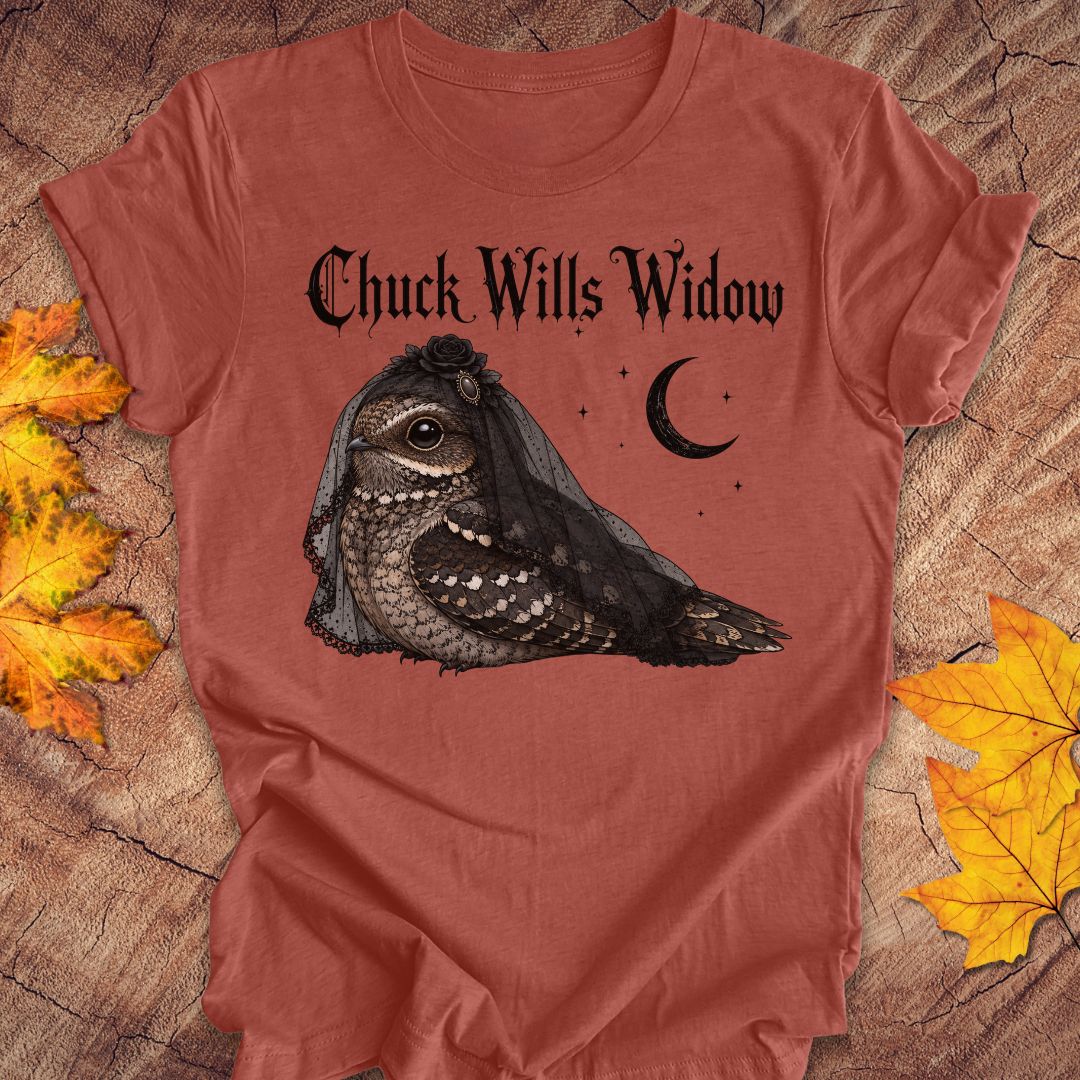 A heathered terracotta t-shirt featuring a gothic-style graphic of a nightjar bird wearing a black widow veil, with the text 'Chuck Wills Widow' and a crescent moon. The t-shirt is styled flat on a rustic wood background with autumn leaves.