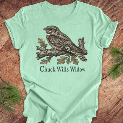 A mint green short-sleeve t-shirt laid flat on a rustic wood surface, featuring a detailed graphic of a brown and gray night hawk on a branch with the text 'Chuck Wills Widow' printed below.