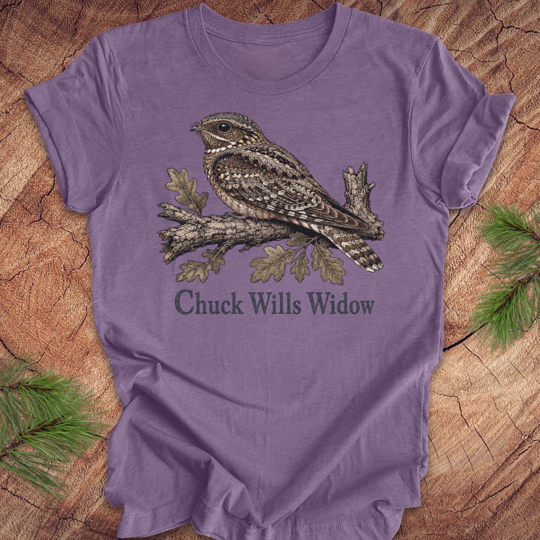 A purple short-sleeve crewneck t-shirt featuring a detailed graphic of a brown and gray camouflage night hawk on a branch with the text 'Chuck Wills Widow' underneath, displayed flat on a rustic wood background with pine needles.