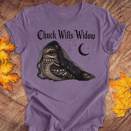 A lavender purple graphic t-shirt featuring a gothic-style illustration of a nightjar bird wearing a black widow veil, with the text 'Chuck Wills Widow' and a crescent moon. The t-shirt is displayed flat on a rustic wood background decorated with autumn maple leaves.