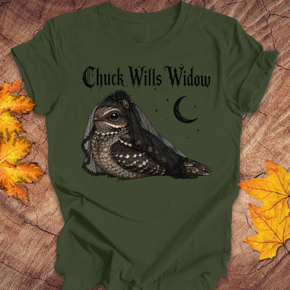 An olive green t-shirt with a graphic of a bird of prey wearing a widow's veil and the text 'Chuck Wills Widow' in a gothic font, displayed on a rustic wood background with autumn leaves.