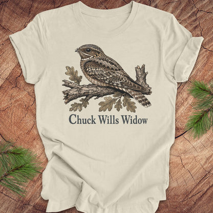 An beige short-sleeve t-shirt with a graphic of a brown and gray night hawk on a branch with the text 'Chuck Wills Widow' underneath, displayed flat on a rustic wood background with pine needles.