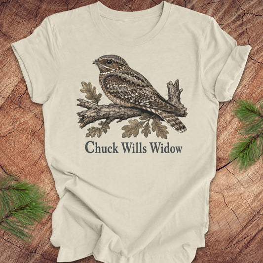 An beige short-sleeve t-shirt with a graphic of a brown and gray night hawk on a branch with the text 'Chuck Wills Widow' underneath, displayed flat on a rustic wood background with pine needles.