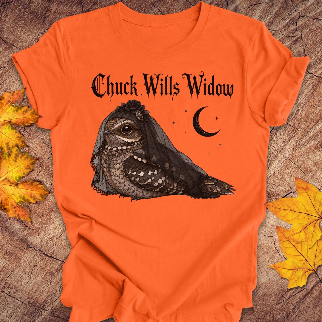 An orange short-sleeve t-shirt laid flat on a rustic wood surface with autumn leaves around it. The shirt features a black graphic of a nightjar owl wearing a widow's veil, with the text 'Chuck Wills Widow' above it and a crescent moon and stars nearby.