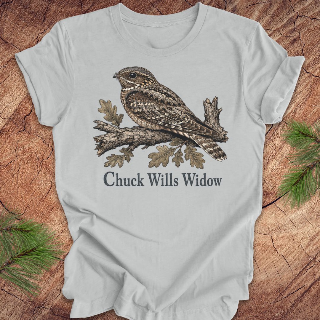 A light gray short-sleeve t-shirt laid flat on a rustic wood background with pine branches in the corners. The shirt features a detailed graphic of a brown and gray camouflage night hawk perched on a branch with the text 'Chuck Wills Widow' printed below.