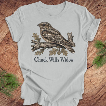 A light gray short-sleeve t-shirt laid flat on a rustic wood background with pine branches in the corners. The shirt features a detailed graphic of a brown and gray camouflage night hawk perched on a branch with the text 'Chuck Wills Widow' printed below.