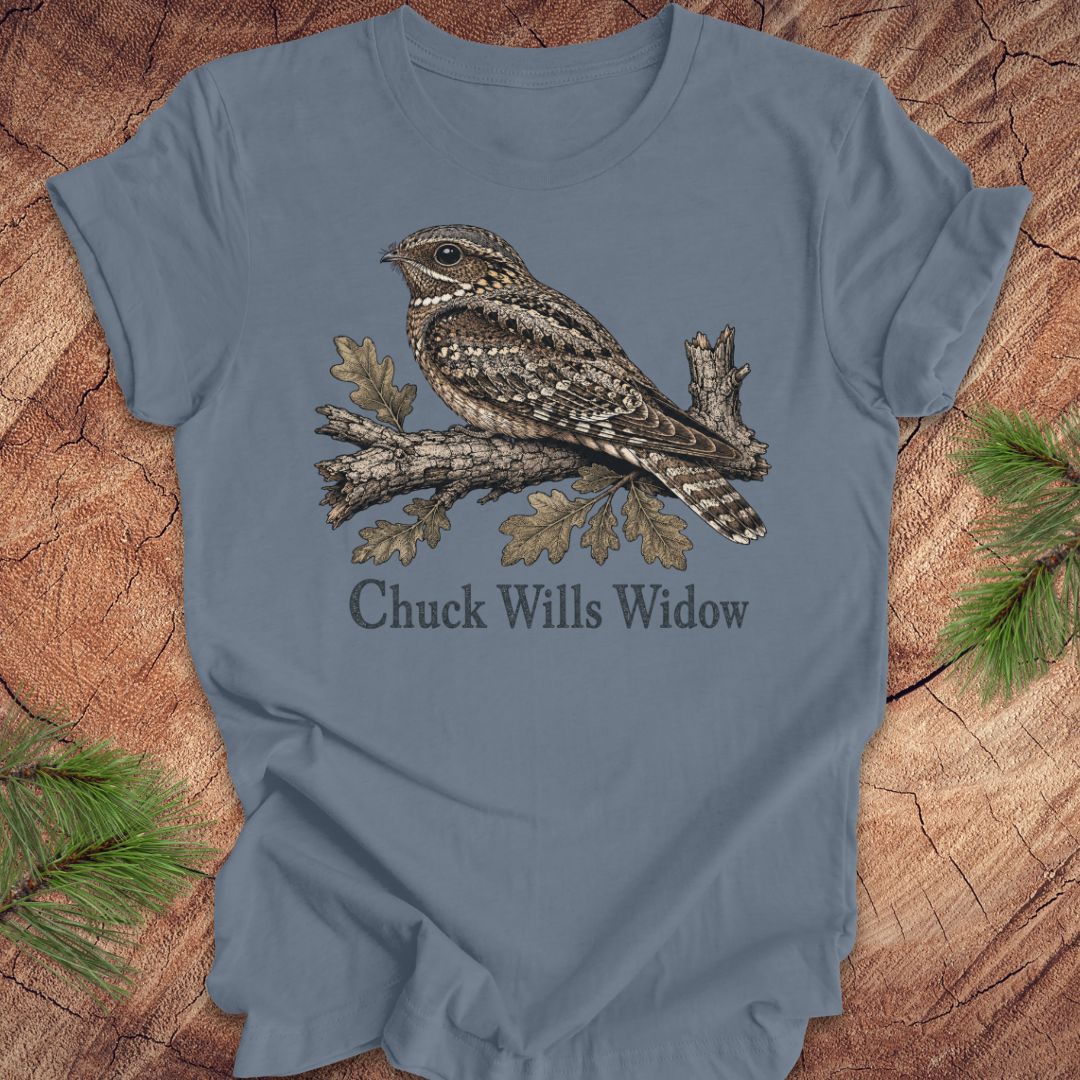 A slate blue short-sleeve t-shirt featuring a detailed graphic of a brown and gray wrybill perched on an oak branch with the text 'Chuck Wills Widow' underneath. The t-shirt is displayed flat on a rustic wood background with pine needles in the corners.