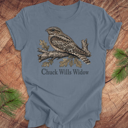 A slate blue short-sleeve t-shirt featuring a detailed graphic of a brown and gray wrybill perched on an oak branch with the text 'Chuck Wills Widow' underneath. The t-shirt is displayed flat on a rustic wood background with pine needles in the corners.