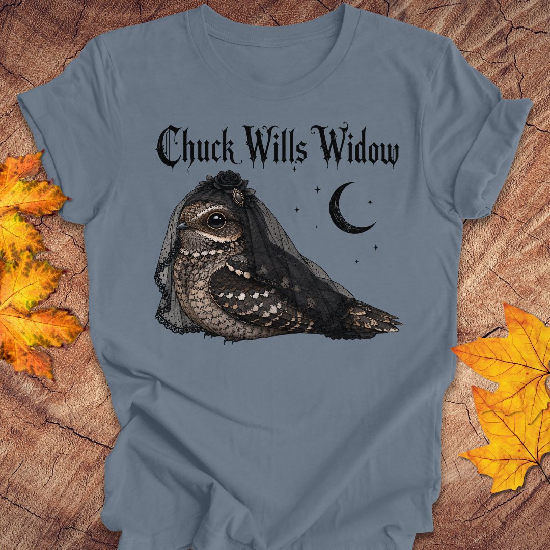 A slate blue crewneck t-shirt featuring a gothic-style graphic of a nightjar bird wearing a widow's veil, with the text 'Chuck Wills Widow' and a crescent moon. The t-shirt is displayed flat on a rustic wood background decorated with autumn leaves.