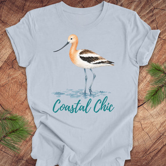 A light blue short-sleeve t-shirt featuring a watercolor illustration of a shorebird and the text 'Coastal Chic' in teal script, laid flat on a rustic wood background with pine branches in the corners.