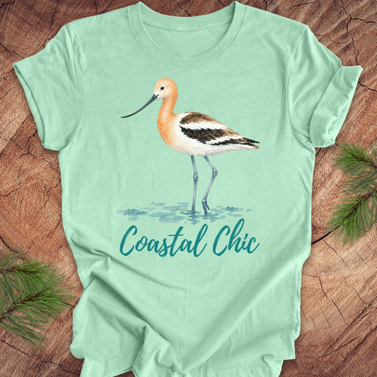 A mint green short-sleeve t-shirt featuring a watercolor illustration of an  wading bird and the text 'Coastal Chic' in teal script, laid flat on a rustic wood background with pine branches.