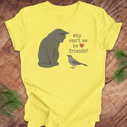 A yellow short-sleeve graphic T-shirt laid flat on a rustic wood surface with pine branches around it. The shirt features a gray cat illustration, a small gray bird, and the text 'why can't we be friends?' with a red heart.