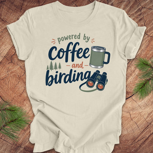 Beige t-shirt with 'powered by coffee and birding' text and graphics on a wooden surface with pine branches.