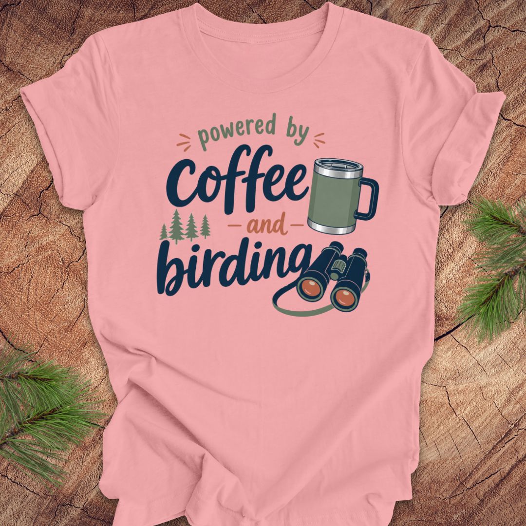 Pink t-shirt with 'powered by coffee and birding' design on a wooden surface with pine branches.