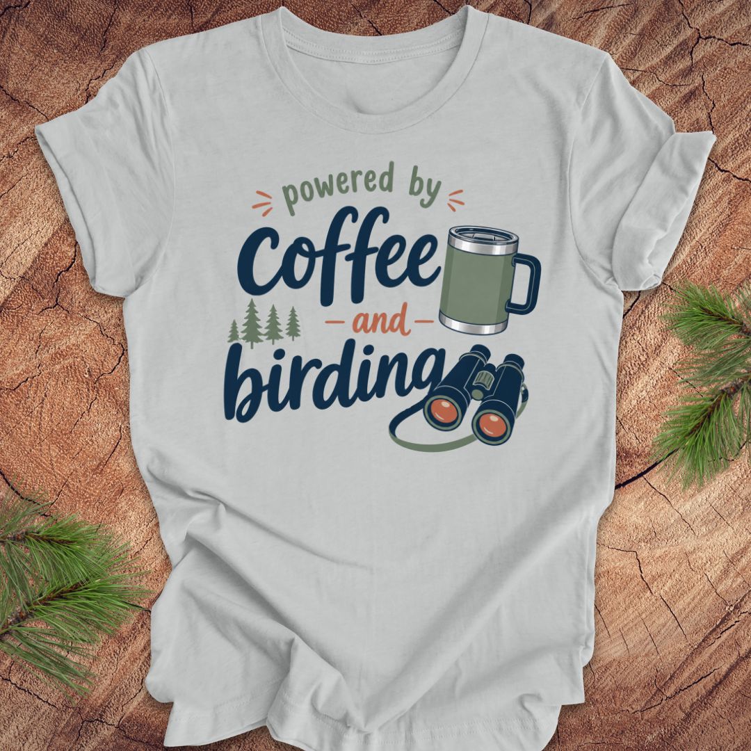 Gray t-shirt with 'powered by coffee and birding' design on a wooden surface with pine branches.