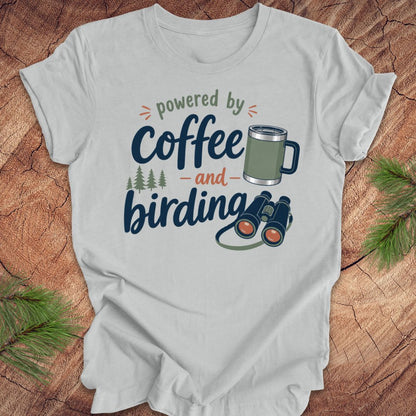Gray t-shirt with 'powered by coffee and birding' design on a wooden surface with pine branches.
