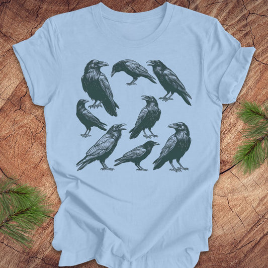 Light blue t-shirt with black crow and raven illustrations on a wooden surface
