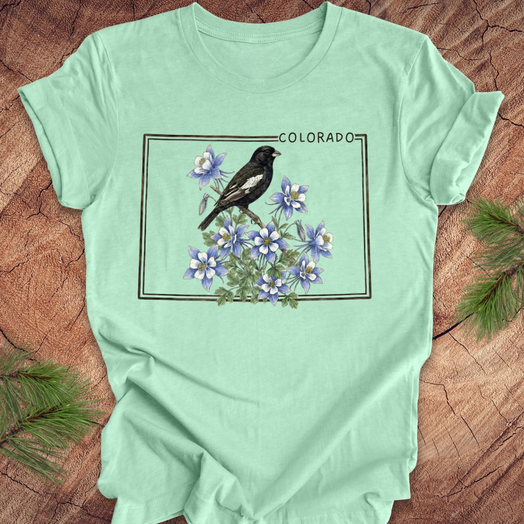 Mint green t-shirt with the outline of colorado with lark bunting and columbine design on a wooden surface