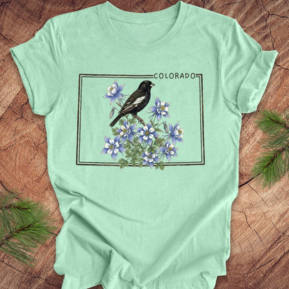 Mint green t-shirt with the outline of colorado with lark bunting and columbine design on a wooden surface