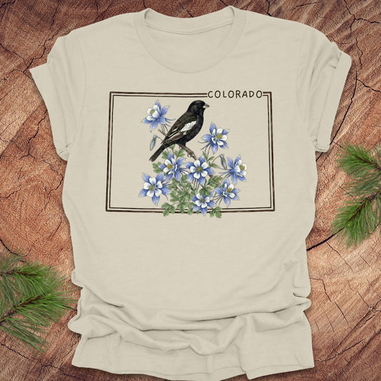 Beige t-shirt with the outline of colorado with lark bunting and columbine design on a wooden surface