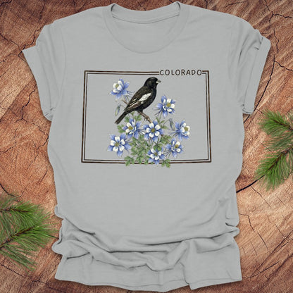 Gray t-shirt with the outline of colorado with lark bunting and columbine design on a wooden surface