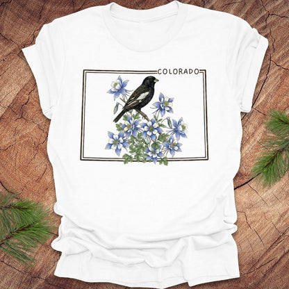 White t-shirt with the outline of colorado with lark bunting and columbine design on a wooden surface