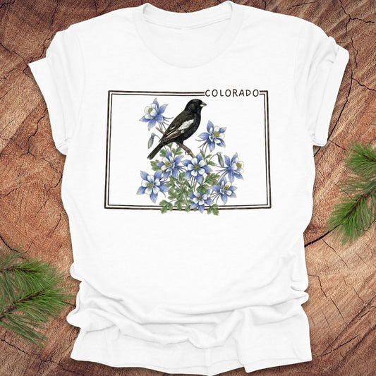White t-shirt with the outline of colorado with lark bunting and columbine design on a wooden surface
