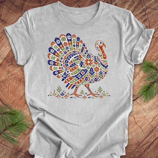 A light gray short-sleeve crewneck t-shirt featuring a colorful, patterned graphic of a Thanksgiving turkey on the chest. The shirt is laid flat on a rustic wood background with pine branches visible in the corners.